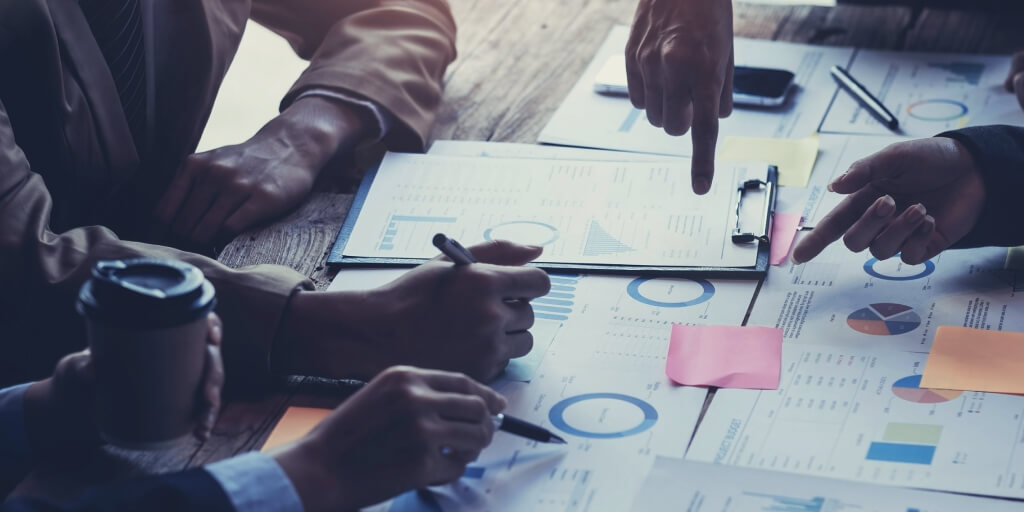 A group of people sitting at the table planning an advertising strategy. Only their hands are in the shot, pointing to different documents laid across the table.