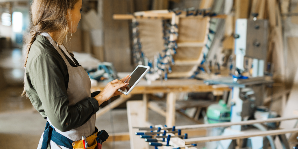 A woman wearing an apron and holding a tablet in a production facility surrounded by unfinished products