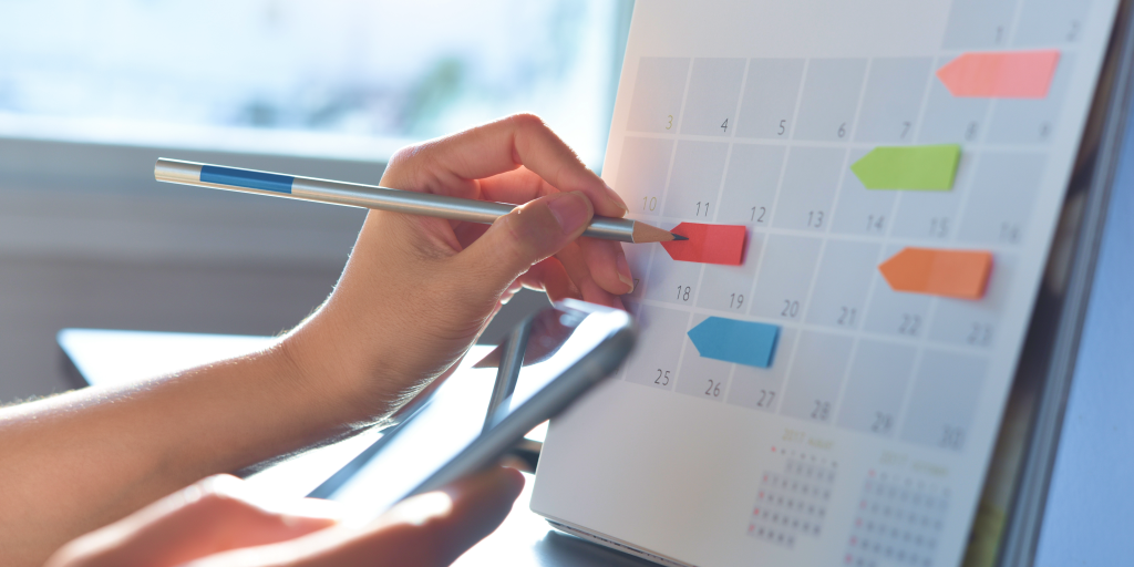 A view of the paper calendar with several colorful marker papers. A hand holding a pencil is writing on the red paper marker. 