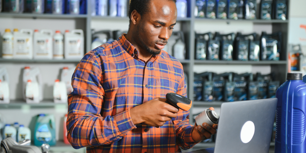 A man standing next to a laptop with a barcode scanner in his left hand and a small car part in the right hand. Various products are on the shelves in the background. 
