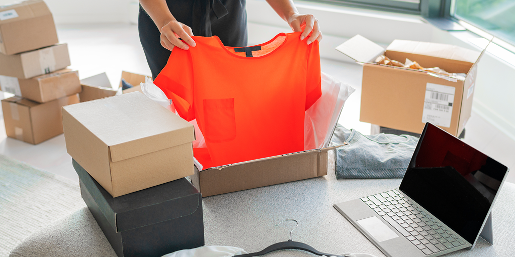 A person is takin an orange t-shirt out of the box. Multiple boxes and a laptop are all around it. 