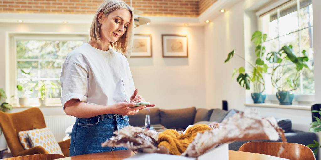 A woman standing in her home. She is holding a smartphone in her hands. 