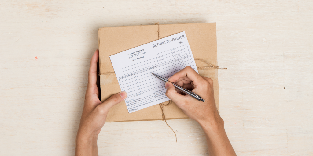 Hands holding a package on a table. A return label is on the top of the package. The person holds a pen in their right hand. 