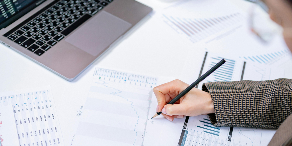 A person writing on a spreadsheet with a pencil. A laptop is in the upper left corner of the picture. 
