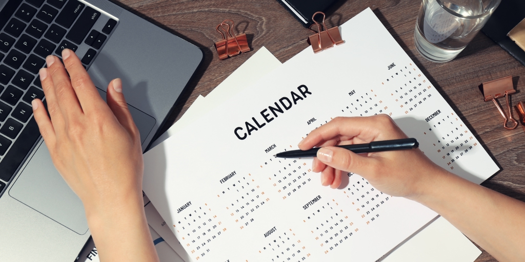 A view of the laptop and a calendar on a desk. The female hand is holding a pen over the calendar and extends her left hand toward the laptop's keyboard.