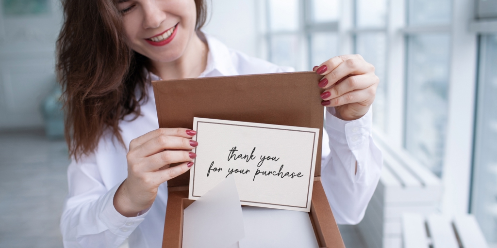 A woman dressed in a white shit is holding a box with a greeting card with the words 'Thank you for your purchase' written on it, 