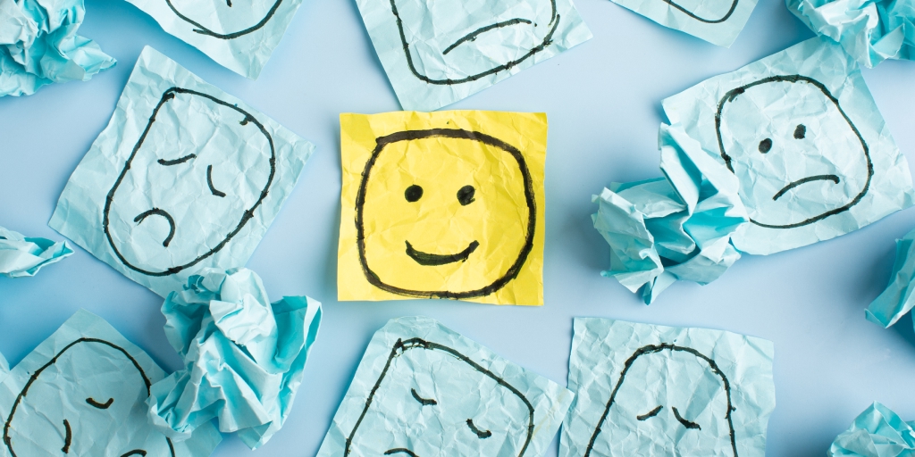 A smiley face on a yellow post-it note. The centrally positioned yellow smiley is surrounded by pale blue crumpled post-it notes with frowny faces drawn on them.