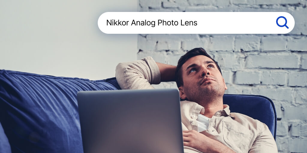 A man laying on a couch with a laptop on his lap. Above his head is a search bar with words Nikkor Analog Photo Lens written inside.